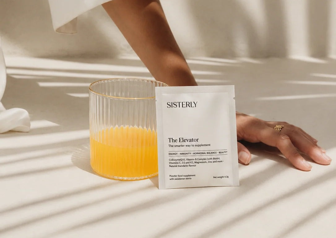 The Elevator sachet beside a glass of the mandarin drink, with a woman's hand resting on the table